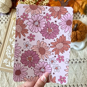 May include: A rectangular floral print card featuring a pattern of pink, mauve, and peach-colored daisies and small stars on a light pink background. The card is held in a hand with a white and pink manicure. The overall theme is floral and decorative.