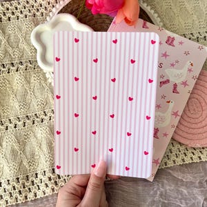 May include: A hand holds a card with pink and white stripes and red hearts. Another card has a pink background with white geese, stars, and cowboy boots. A white, cloud-shaped dish and pink flowers are in the background.