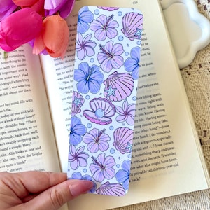 May include: A rectangular bookmark featuring a repeating pattern of purple and light blue hibiscus flowers, seashells, starfish, and pearls on a white background. The design includes small blue dots scattered throughout the floral and ocean theme.