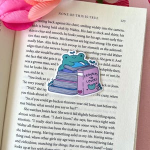May include: A colorful sticker featuring a cartoon frog sitting atop a stack of books. The book on top is titled "Froggies to Lovers." The sticker is placed on an open book with text. Pink and green floral accents are visible.