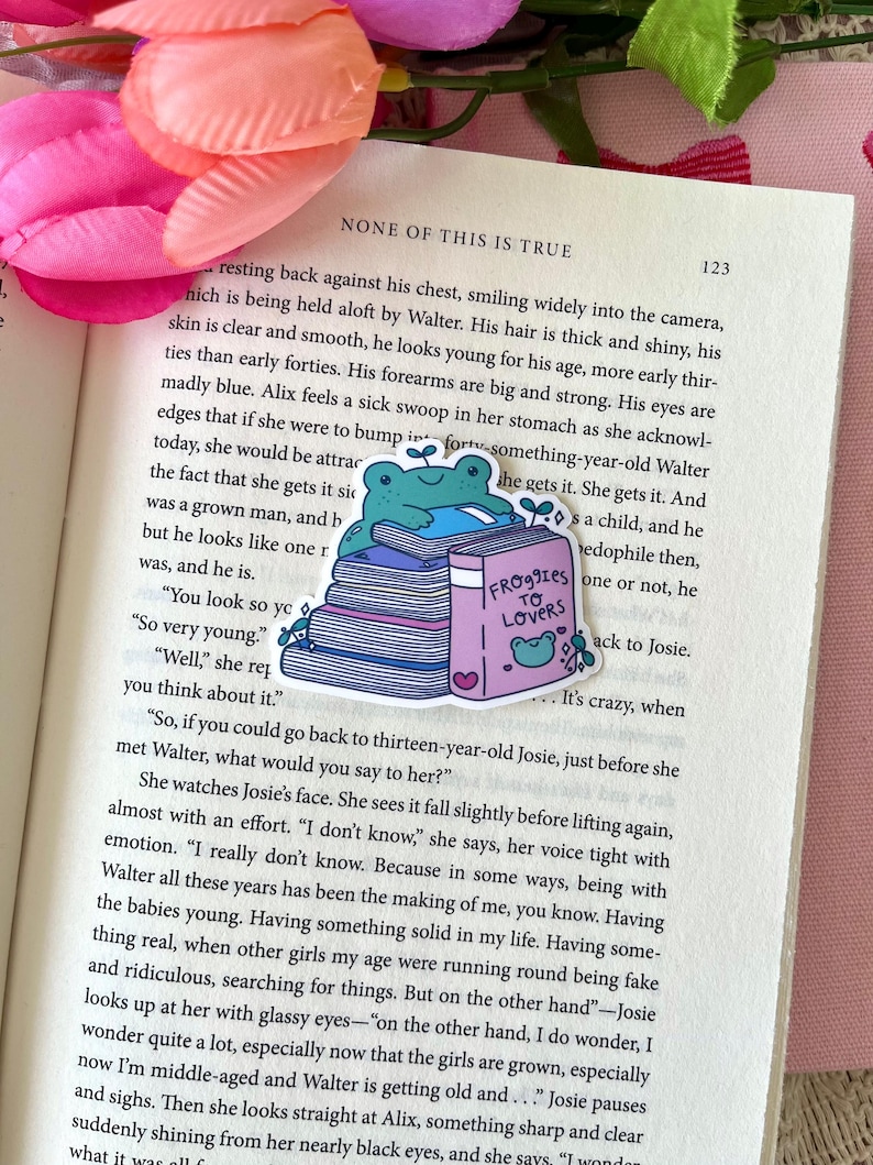 May include: A colorful sticker featuring a cartoon frog sitting atop a stack of books. The book on top is titled "Froggies to Lovers." The sticker is placed on an open book with text. Pink and green floral accents are visible.