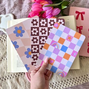 May include: Three colorful patterned bookmarks are displayed, featuring floral, checkerboard, and rainbow designs. The bookmarks are held over an open book, with pink tulips and a pink book with a bow in the background. The color palette includes pink, blue, orange, and brown.