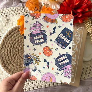 May include: A rectangular card featuring a repeating pattern of Halloween-themed illustrations including orange pumpkins, purple cauldrons with the text Hocus Pocus, a blue book labelled Magic, crystal balls, bats, and stars on a light background.