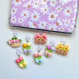 May include: A clear phone case with a daisy pattern and various decorative charms. The charms feature miniature flower arrangements, including tulips and roses, with bows and a small picket fence.