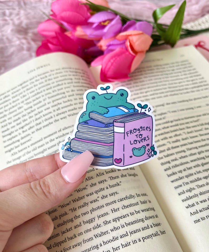 May include: A sticker featuring a teal frog sitting atop a stack of books. The book on top has the title "Froggies to Lovers" and a heart. The sticker is on a book with pink flowers in the background.