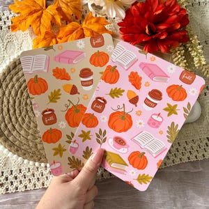 May include: Two rectangular cards featuring a repeating autumn-themed pattern. One card has a beige background, the other a light pink background. Both display illustrations of orange pumpkins, open books, coffee cups, candles labelled fall, and leaves.