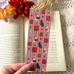 May include: A rectangular bookmark with a pink background featuring vertical stripes and a repeating pattern of ghosts reading books, black cats, orange jack-o'-lanterns, red hearts, skulls, spiderwebs, and red books labelled with the text BOOK LOVER.