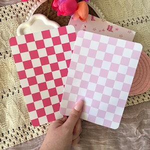 May include: Two rectangular pink and white checkered notepads. One notepad is held by a hand. A pink and white checkered pattern is on the front of the notepad. A pink and white checkered pattern is on the other notepad. Pink tulips are in the background.