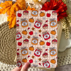 May include: A rectangular card featuring a repeating pattern of orange pumpkins, red apples, baskets of apples, and jars labelled pumpkin butter or apple jam on a beige gingham background. Small white flowers and brown autumn leaves are scattered throughout.