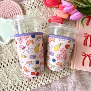 May include: Two clear plastic cups with purple and white patterned fabric sleeves. The sleeves feature a pink background with illustrations of lemons, cherries, croissants, teacups, and stars.