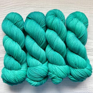 Op de afbeelding: Vier strengen fel turquoise garen die naast elkaar zijn gerangschikt. Elke streng is strak gedraaid in een uniforme, cilindrische vorm, met een consistente, gladde textuur en een heldere, verzadigde kleur over de gehele vezel.
