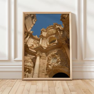Può includere: Fotografia incorniciata di una facciata di cattedrale ornata, ripresa dal basso. La struttura in pietra beige presenta intagli dettagliati, colonne e archi contro un cielo blu limpido. La cornice in legno completa i toni caldi dell'edificio.