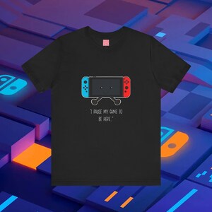 May include: Black t-shirt with a white outline of a Nintendo Switch console with red and blue joy-cons. The text "I pause my game to be here." is printed below the console.
