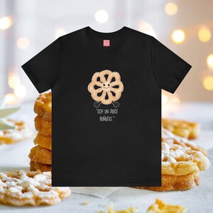 May include: Black t-shirt featuring a cartoon bunuelo character with a smiling face and the text "SOY UN DULCE BUÑUELO." The shirt has a small red tag on the upper back. The background shows a stack of bunuelos.