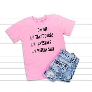 Op de afbeelding: Roze T-shirt met de tekst "Dag vrij: Tarotkaarten, Kristallen, Heksenshit" met vinkjes naast elk item op de lijst. Het shirt wordt gecombineerd met een spijkerbroek.