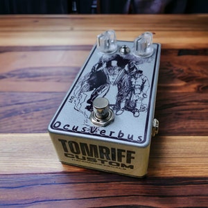 Op de afbeelding: Een zilveren gitaareffectpedaal met een zwart-witte illustratie van twee figuren. Het pedaal is gelabeld "Ocus-Verbus" en "Tomriff Custom Handmade Pedals".