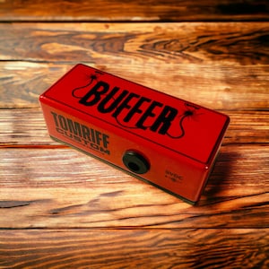 May include: A red Tomriff Custom Buffer pedal with the word "BUFFER" in black lettering and a black power jack. The pedal has a black label with the text "9VDC" and a black label with the text "Tomriff Custom Handmade Pedals".