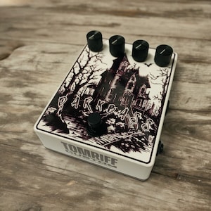 Könnte beinhalten: Weißes Gitarreneffektpedal mit schwarzen Knöpfen und einer Schwarzweißillustration eines Spukhauses mit dem Text "Darkbass" darauf. Das Pedal ist mit "Tomriff Custom" beschriftet.