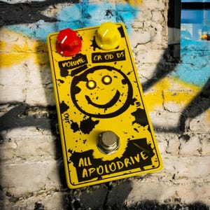 Könnte beinhalten: Gelbes Gitarrenpedal mit einem Smiley und dem Text "ALL APOLODRIVE" in Schwarz. Das Pedal hat drei Knöpfe mit den Beschriftungen "VOLUME", "CR OD DS" und einen schwarzen Fußschalter.