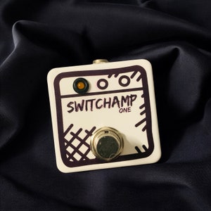 Puede incluir: Pedal de guitarra blanco con marcas negras y moradas. El pedal tiene un botón grande en el centro y dos botones más pequeños en la parte superior. El texto "SWITCHAMP ONE" está impreso en el pedal.