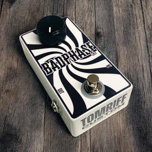 Könnte beinhalten: Ein weißes Gitarreneffektpedal mit einem schwarz-weißen Wirbeldesign und dem Text "BADPHASE 45". Es hat einen schwarzen Knopf, eine silberne Ein-/Ausgangsbuchse und einen goldfarbenen Fußschalter. Der Text "TOMRIFF CUSTOM" ist ebenfalls sichtbar.