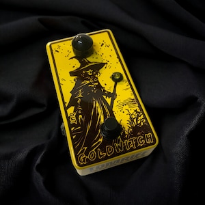 Peut inclure: Une pédale d'effets de guitare jaune avec une illustration en noir et blanc d'une sorcière tenant un bâton. La pédale porte l'inscription "Goldwitch".