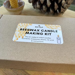 May include: A brown cardboard box with a white label that says "Beeswax Candle Making Kit". The label has a honeycomb pattern and a small illustration of a bee. The text on the label says "This kit makes 6 rolled candles. Contents: 6 beeswax sheets, 1m wick & instructions."