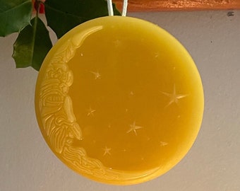 May include: A yellow, round, wax ornament with a crescent moon and stars design. The ornament is hanging from a white string.
