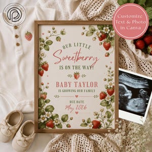 May include: A rectangular wooden frame containing a pregnancy announcement sign with strawberry illustrations, green leaves, and white flowers. The text reads: Our Little Sweetberry Is On The Way! Baby Taylor Is Growing Our Family Due Date May 2026.