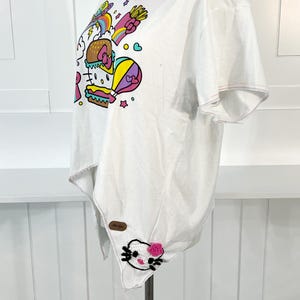 May include: White t-shirt featuring a colorful graphic of a burger, fries, and a hot air balloon with a cartoon character. The shirt has a unique asymmetrical hemline and a pink crocheted collar. A small patch with a cartoon character is sewn on the hem.