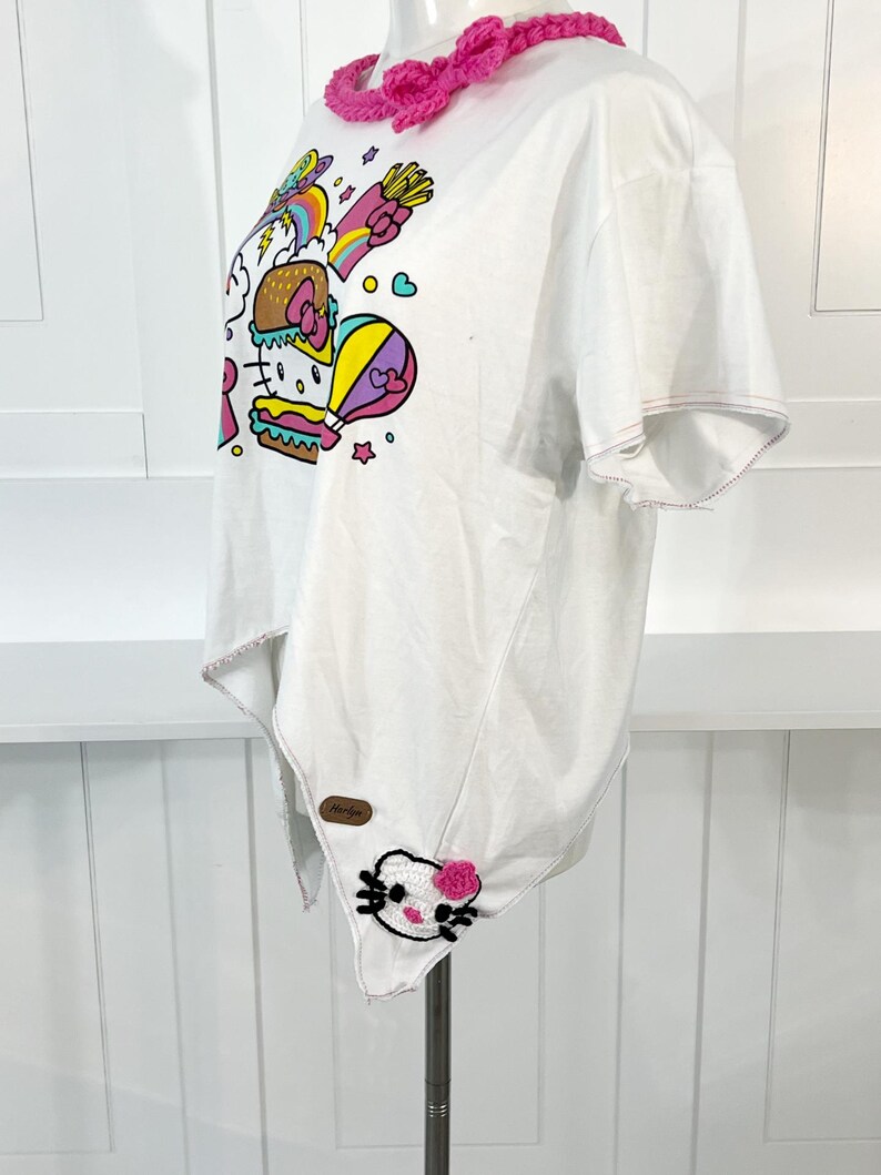 May include: White t-shirt featuring a colorful graphic of a burger, fries, and a hot air balloon with a cartoon character. The shirt has a unique asymmetrical hemline and a pink crocheted collar. A small patch with a cartoon character is sewn on the hem.