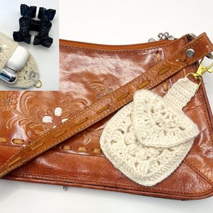 May include: A brown leather purse with a floral design and a cream-colored crochet pouch attached. The pouch is square with a flap and a small strap. A black decorative object and a crochet case with earbuds are in the upper left corner.