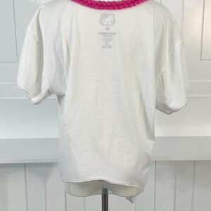 May include: White t-shirt with a pink, braided collar. The shirt features a small Hello Kitty graphic near the collar. The back of the shirt is visible, showing the neckline and the bottom hem.