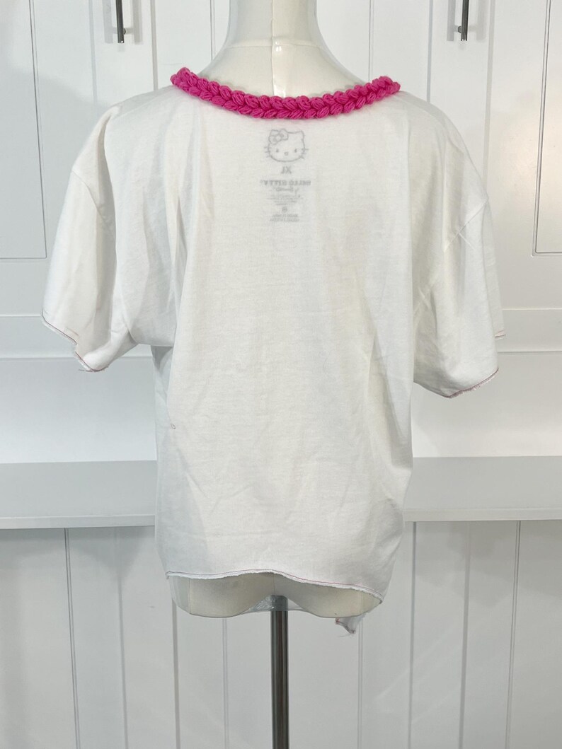 May include: White t-shirt with a pink, braided collar. The shirt features a small Hello Kitty graphic near the collar. The back of the shirt is visible, showing the neckline and the bottom hem.