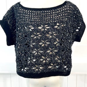 May include: A black and silver crochet top with short sleeves. The top features a scoop neckline and a decorative openwork pattern. The edges of the top and sleeves are finished with a solid black trim. The crochet design creates a semi-sheer effect.