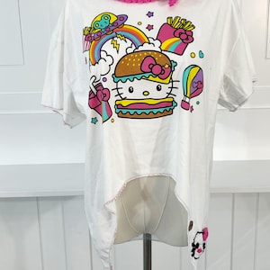 May include: White t-shirt featuring a colorful graphic of a Hello Kitty burger, rainbow, and UFO. The shirt has a pink crocheted neckline and a unique asymmetrical hemline. The edges of the shirt are finished with pink stitching.