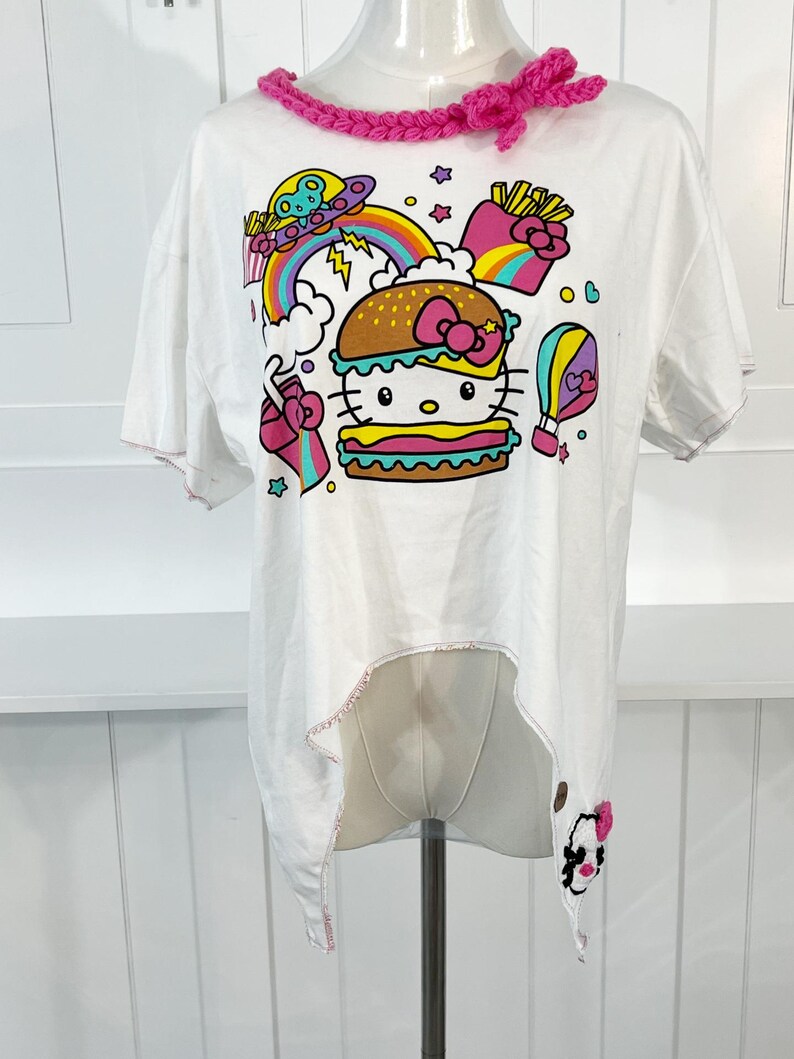 May include: White t-shirt featuring a colorful graphic of a Hello Kitty burger, rainbow, and UFO. The shirt has a pink crocheted neckline and a unique asymmetrical hemline. The edges of the shirt are finished with pink stitching.