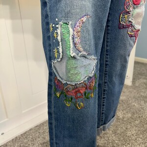 May include: Blue denim jeans with embroidered patches. The patches feature colorful floral and celestial designs, including a crescent moon and stars. The jeans have a distressed look with rolled-up cuffs.