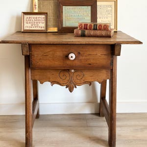 May include: A small, antique wooden table with a single drawer and decorative carved apron. The drawer has a white and patterned knob. Framed vintage alphabet samplers and books rest on the table's surface. The table is approximately 30 inches tall.