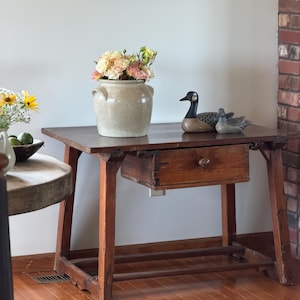 May include: A wooden side table with a single drawer and a central knob, featuring four legs connected by a lower stretcher. On the table sits a beige ceramic crock filled with light-colored flowers and two decorative duck figurines.
