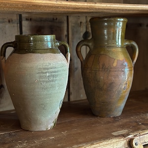 May include: Two antique ceramic jars with green and brown glaze sit on a wooden shelf. The jars have a rounded body and two handles.