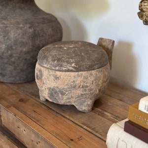 May include: A dark brown, round, ceramic stool with a lid. The stool has a textured surface and is sitting on a wooden table.