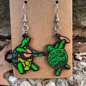 Puede incluir: Un par de pendientes de tortuga verde y marrón con ganchos plateados. Las tortugas están pintadas con pintura verde, marrón y amarilla. Los pendientes están colgados de una tarjeta marrón.