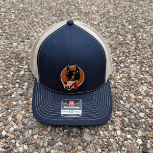 May include: Navy blue and tan trucker hat with a patch featuring a cartoon wolf. The hat has a curved brim and an adjustable strap. The patch has the text "R.R. 1876" and "RICHARDSON ORIGINAL 112 TRUCKER ADJUSTABLE".