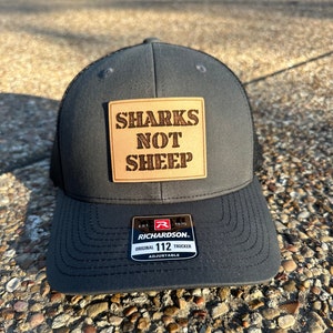 May include: A dark gray trucker hat with a black mesh back. The hat features a rectangular patch with the words "SHARKS NOT SHEEP" in a brown, rustic font. The hat is adjustable and has a Richardson 112 tag.