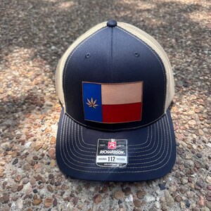 May include: Navy blue trucker hat with a tan mesh back and a curved brim. The front features a leather patch with the Texas flag design, including a cannabis leaf. The hat has the Richardson 112 Trucker label.