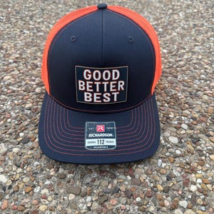 Op de afbeelding: Marineblauwe en oranje truckerpet met de tekst "GOOD BETTER BEST" op een rechthoekige patch. De pet heeft een mesh achterkant en een gebogen klep. Het label van het merk Richardson is zichtbaar.
