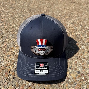Puede incluir: Gorra de camionero azul marino con la parte trasera de malla gris. La parte delantera presenta un diseño de calavera con un sombrero de copa rojo, blanco y azul. La gorra tiene la etiqueta de la marca Richardson y es ajustable.