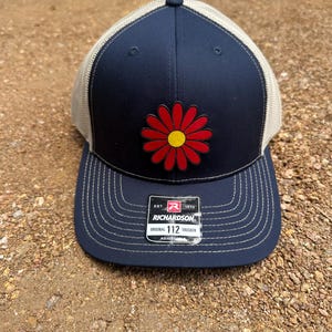 May include: Navy blue trucker hat featuring a red and yellow daisy patch. The hat has a beige mesh back and a curved brim with decorative stitching. The tag reads "RICHARDSON ORIGINAL 112 TRUCKER ADJUSTABLE".