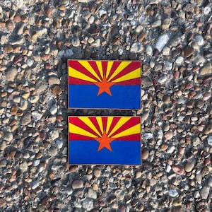 May include: Two rectangular wooden plaques featuring the Arizona state flag design. The flag has a blue field with a copper star, a red and yellow sunburst, and a gold border. The plaques are displayed on a textured, stone surface.